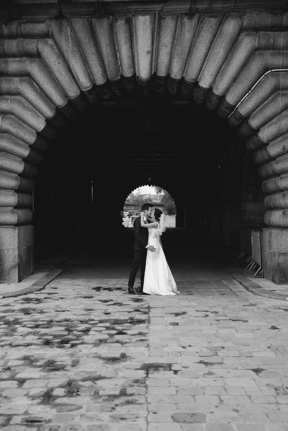 Black and white wedding photo of bride and groom kissing under a grand stone arch on cobblestone pavement.