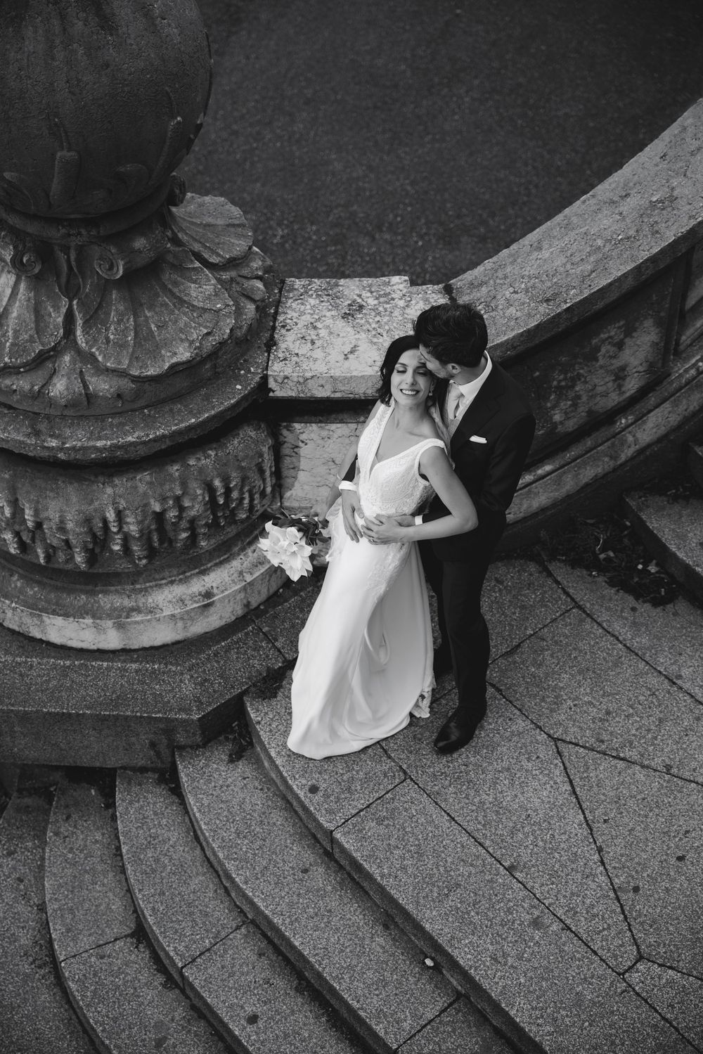 Aerial black and white photo of bride and groom embracing on ornate stone steps with bouquet.