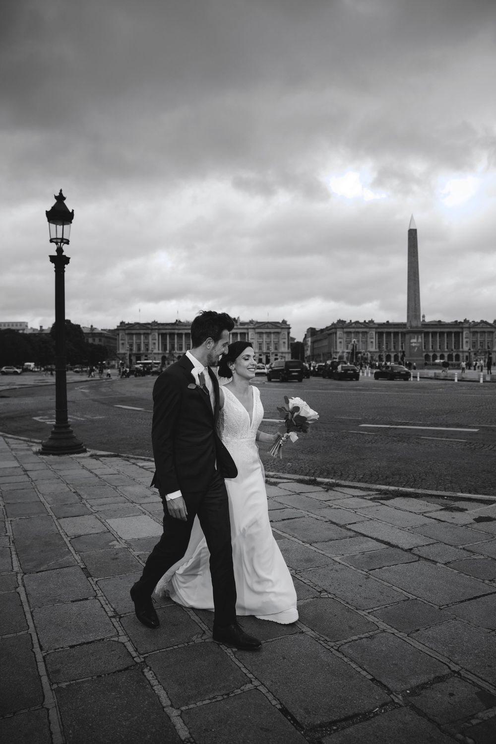 Black and white photo of a bride and groom walking at Place de la Concorde in Paris with the Luxor Obelisk in background.