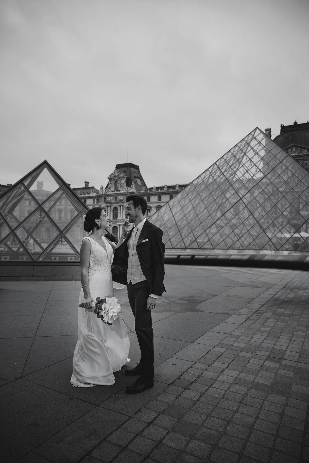 Bride and groom pose romantically before the Louvre Pyramid in Paris in a black and white wedding photo.