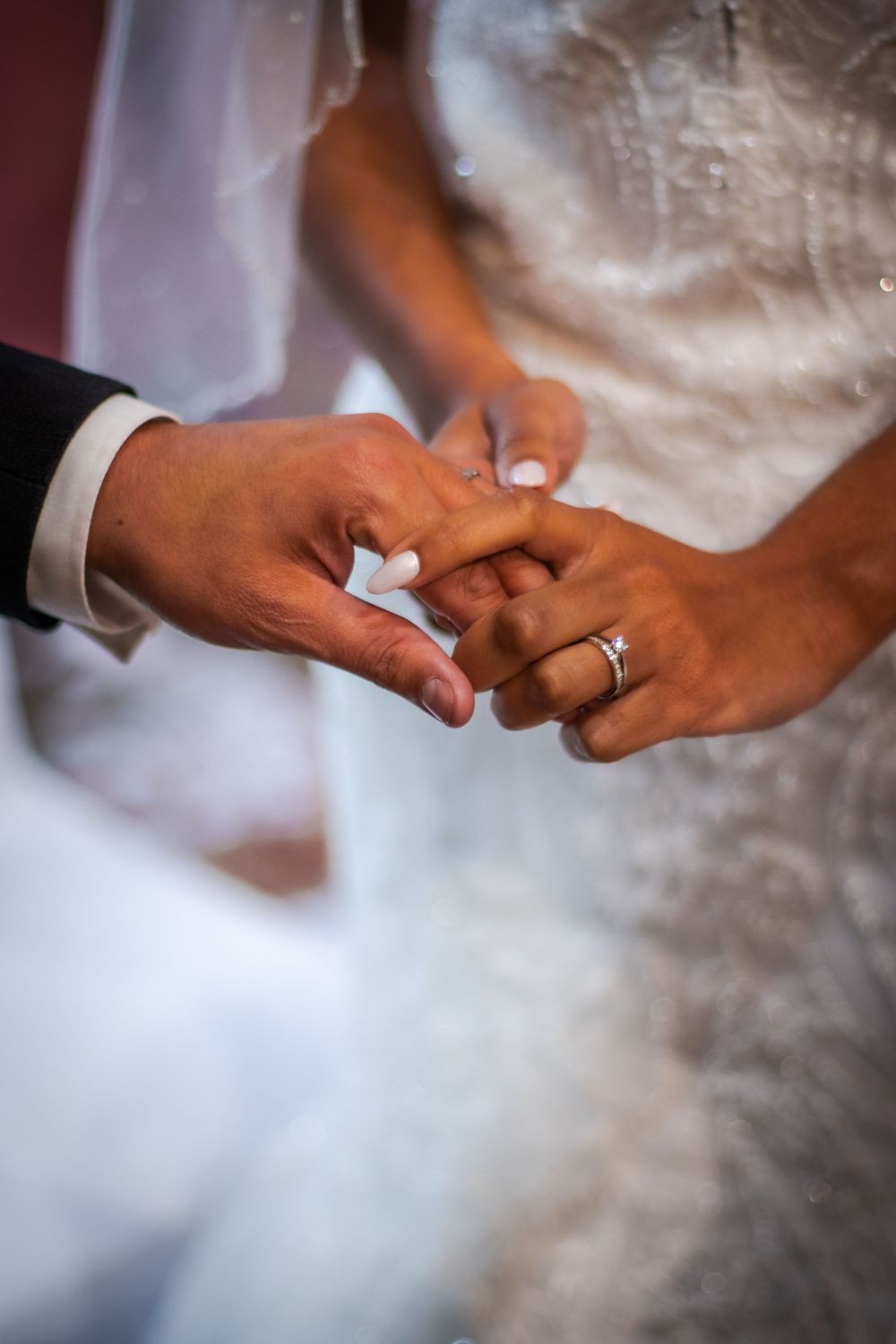 Close up of hands exchanging wedding rings during ceremony with white dress visible in background.