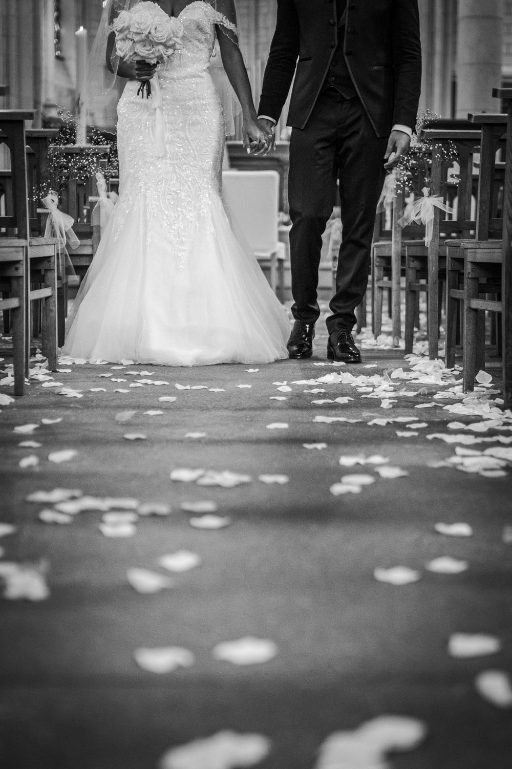 Black and white panoramic image of flower petals scattered on church floor with wedding dress and formal attire visible.
