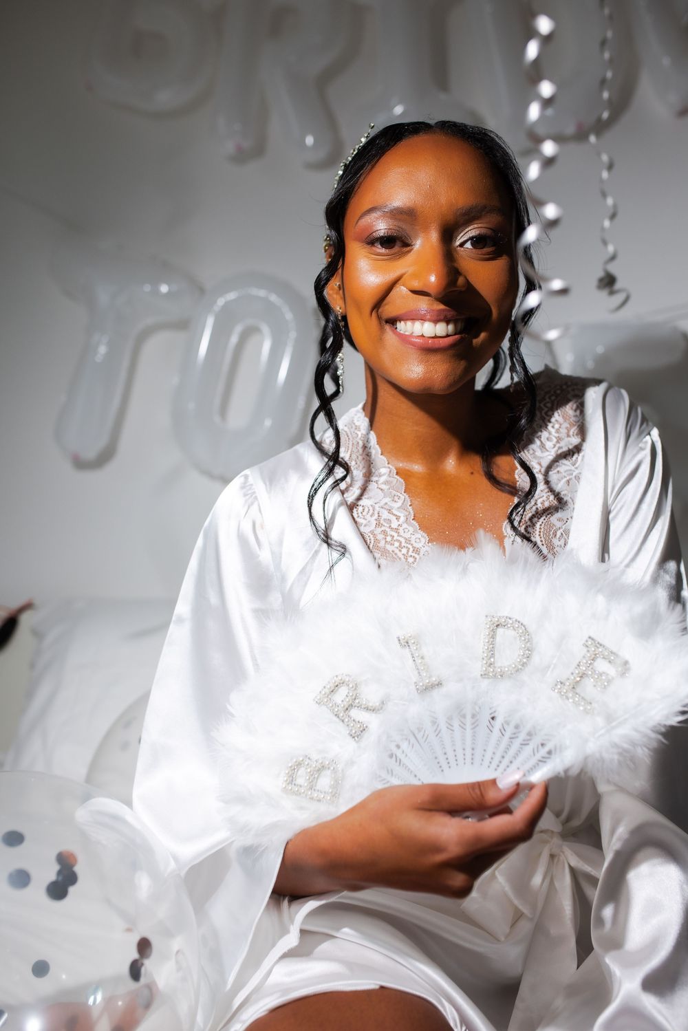 A bride wearing a white satin robe with lace trim holds a decorative white fan against a light background.