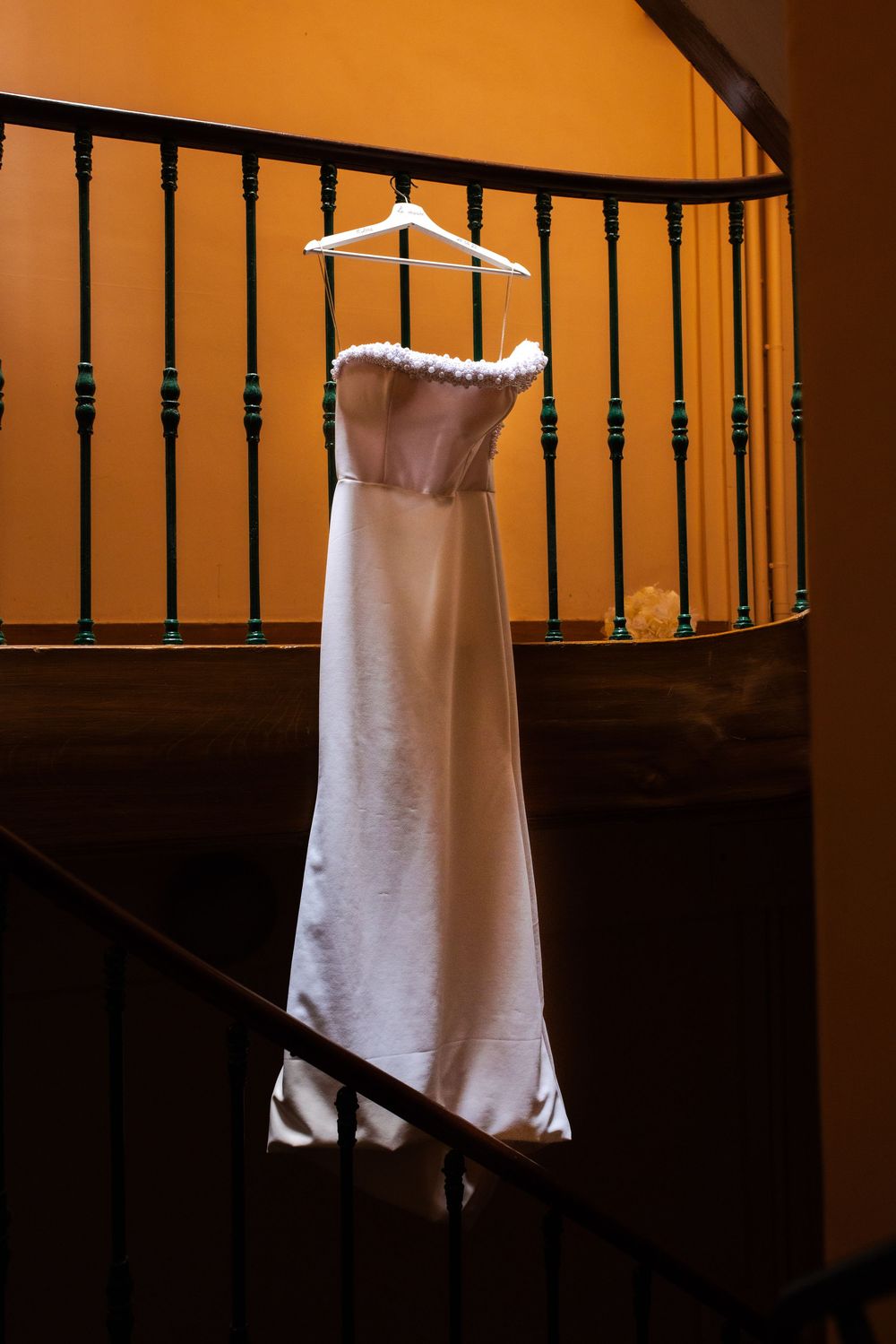A white dress hangs from dark wooden railing bars in dramatic lighting creating contrast between shadows and highlights.