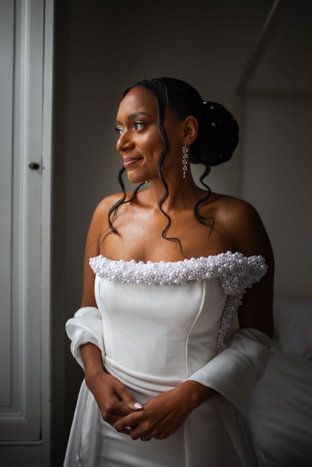 Beautiful bride in off-shoulder white wedding dress poses elegantly in natural window light.