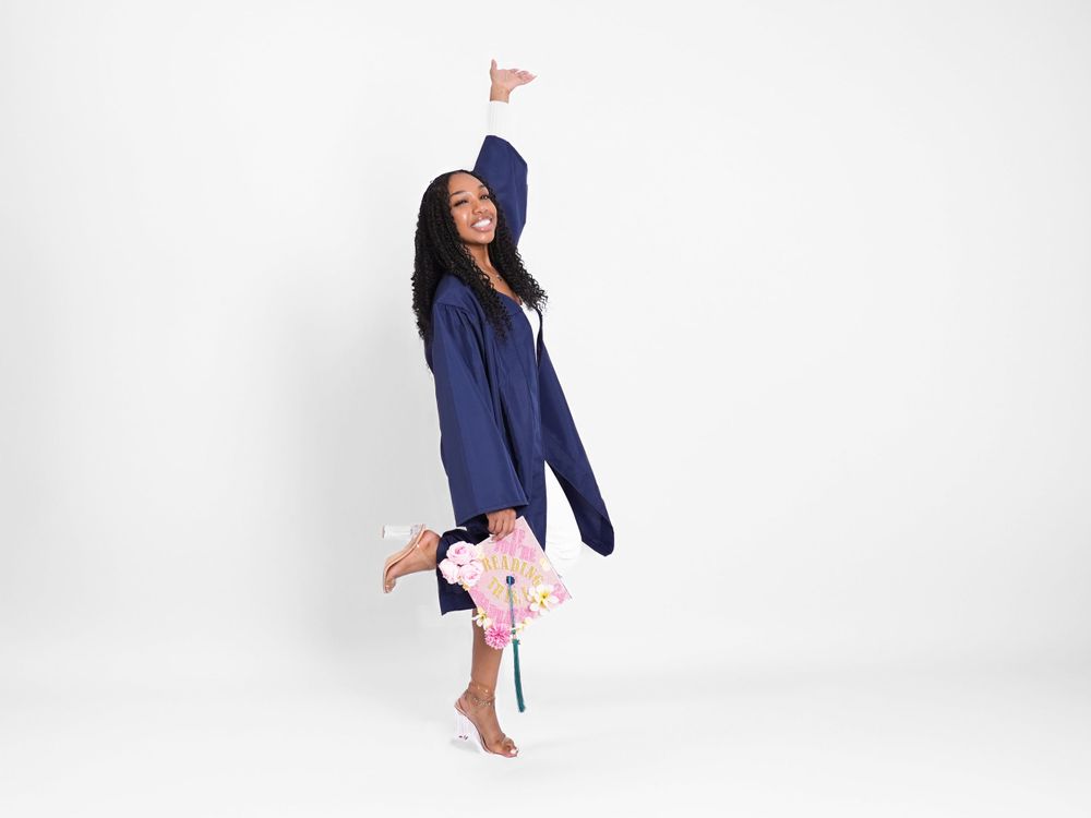 Joyful person in navy blue outfit jumps playfully against white background holding pink flowers.