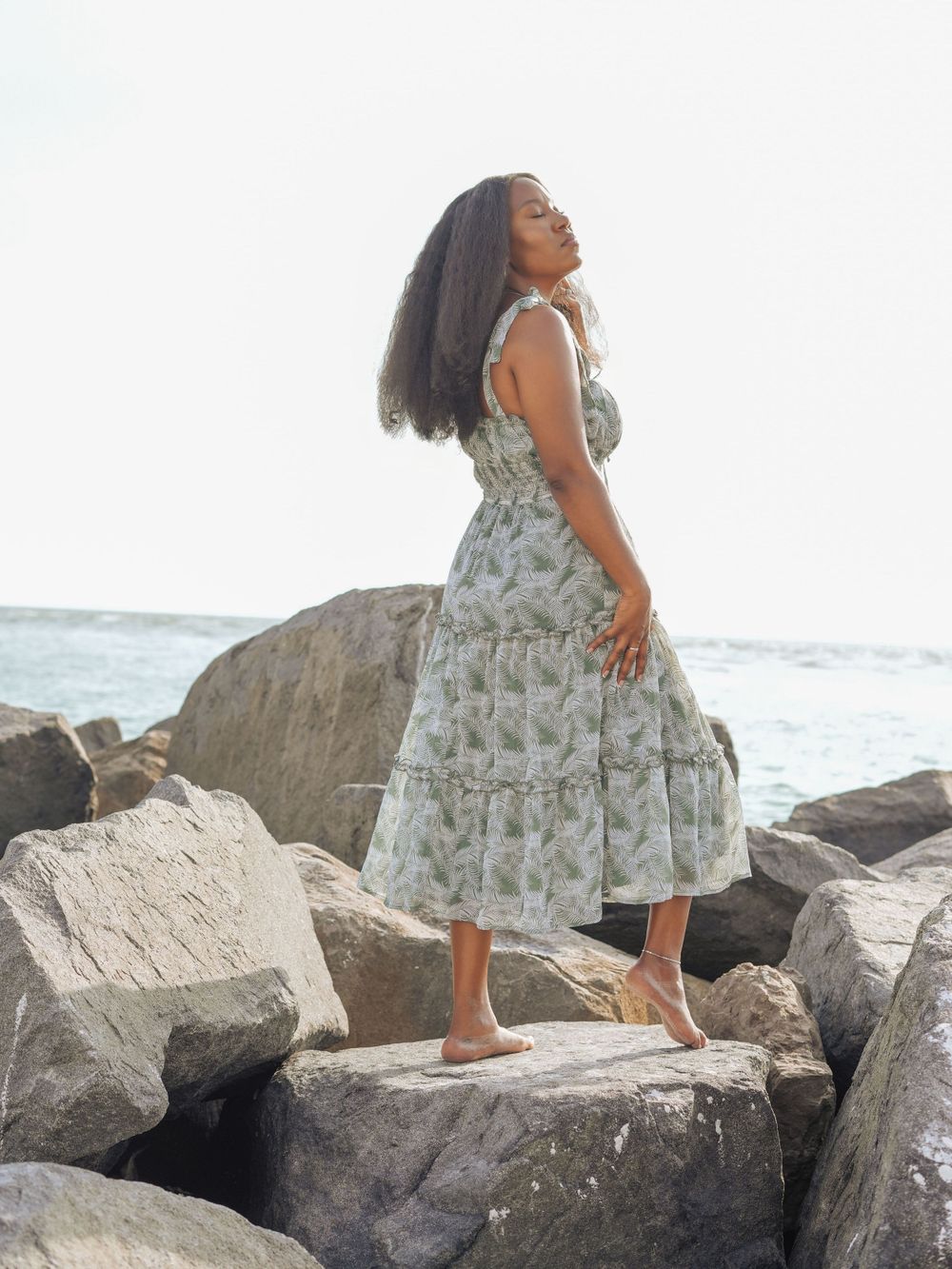Person in floral dress poses dramatically on coastal rocks by the ocean.
