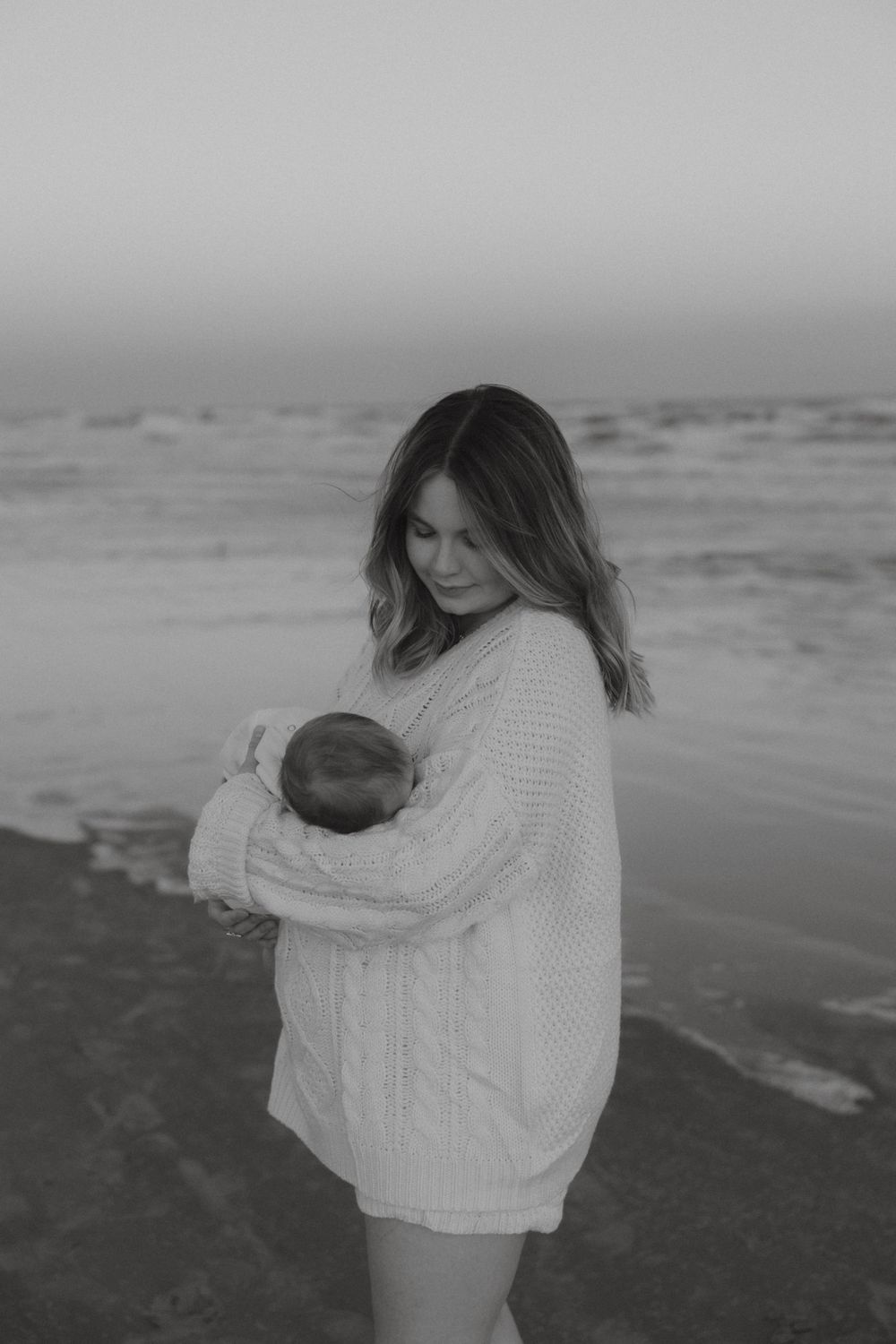 Black and white portrait of mother gazing lovingly at newborn baby on ocean shoreline.