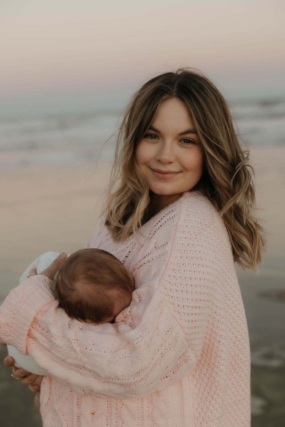 Smiling mother in pink knit sweater cuddling newborn baby on beach at sunset.