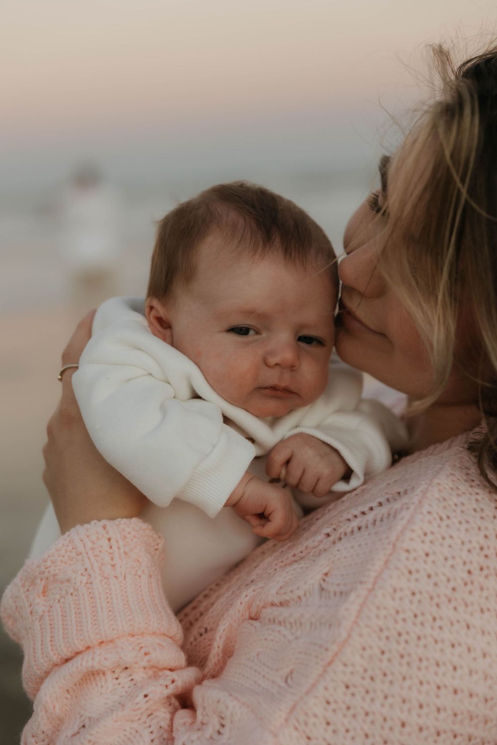 Mother kissing newborn baby on cheek while holding infant against pink knit sweater at beach.