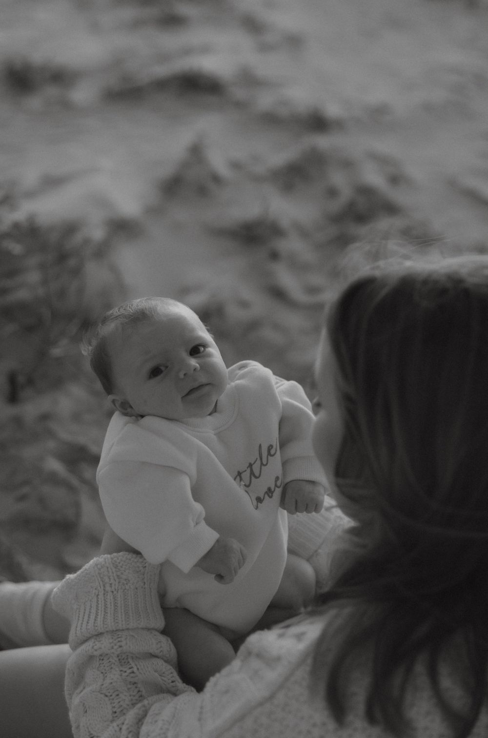 Black and white photo of mother holding newborn baby on sandy beach.