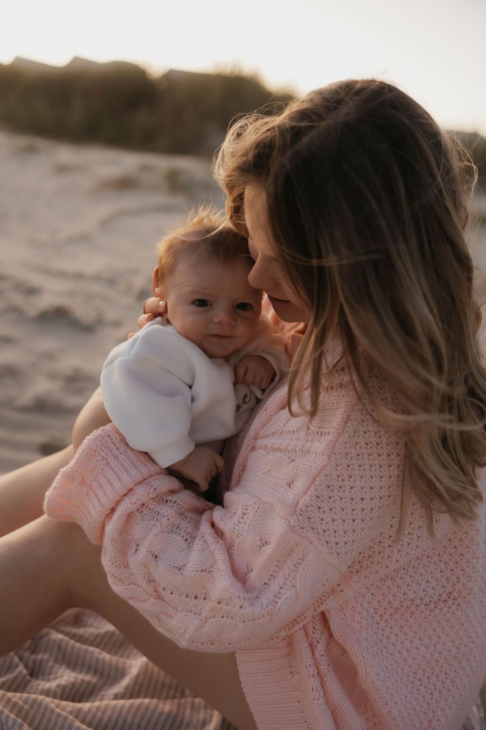 Mother in pink knit sweater holds newborn baby close on sandy beach at sunset.