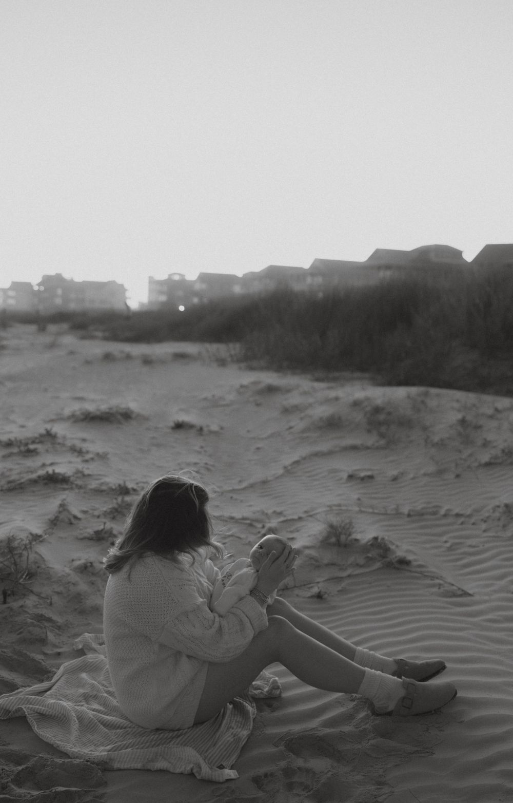 Woman sitting alone on sandy beach dunes at dusk in black and white photography.