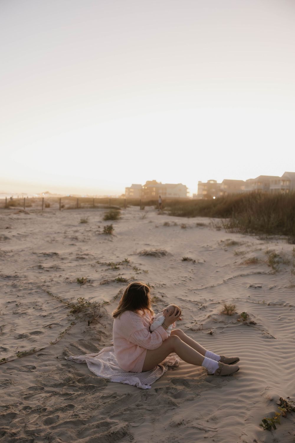 Mother in pink sweater sits on sandy beach holding newborn baby at golden sunset.