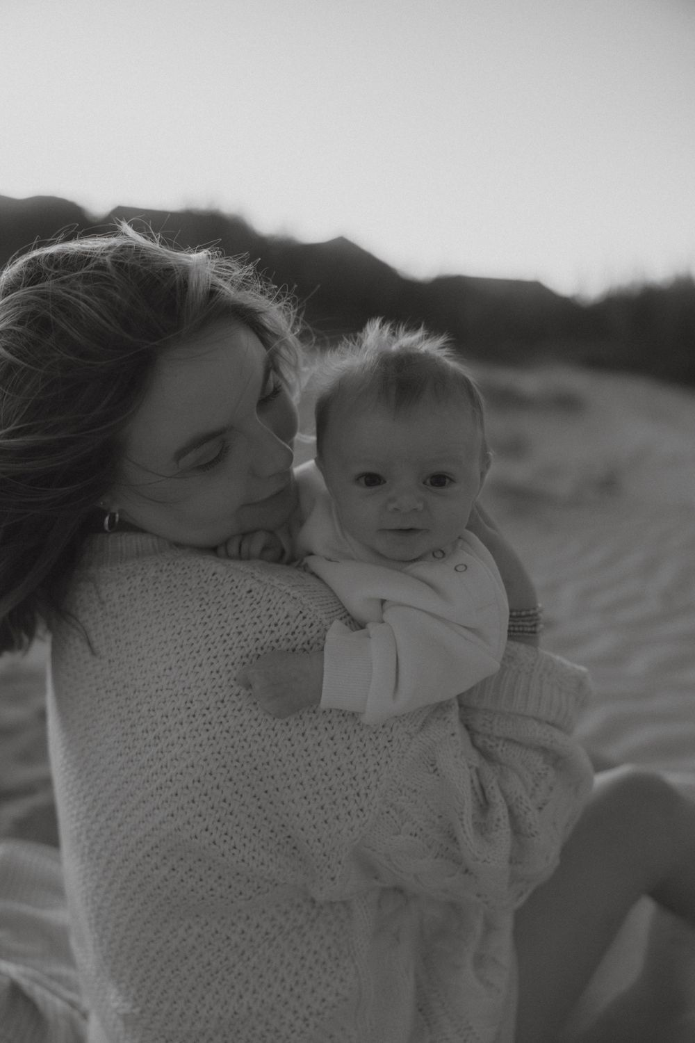 Black and white portrait of mother tenderly holding newborn baby on sandy beach.