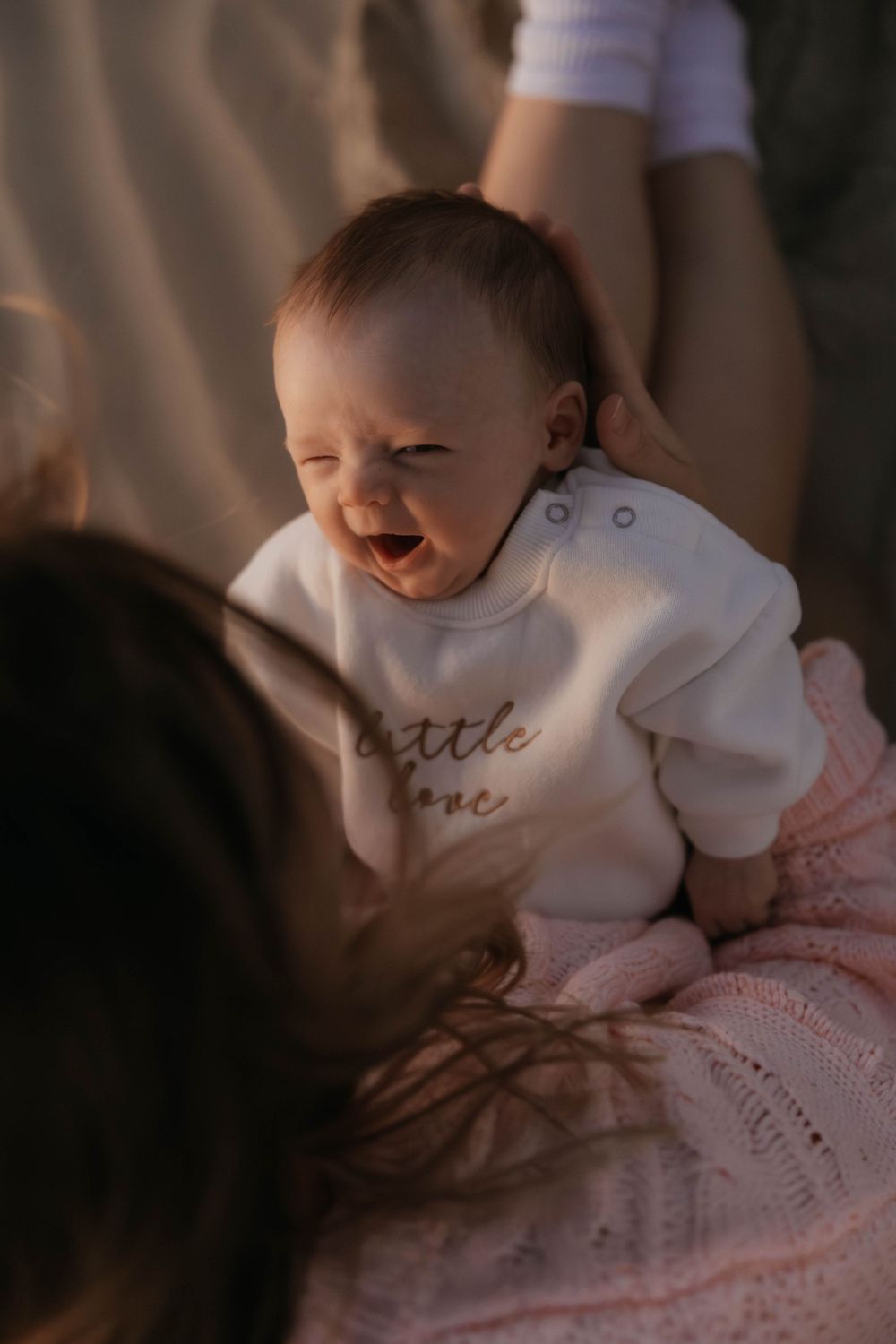 Adorable laughing baby in white 'little love' onesie lying on pink blanket outdoors.