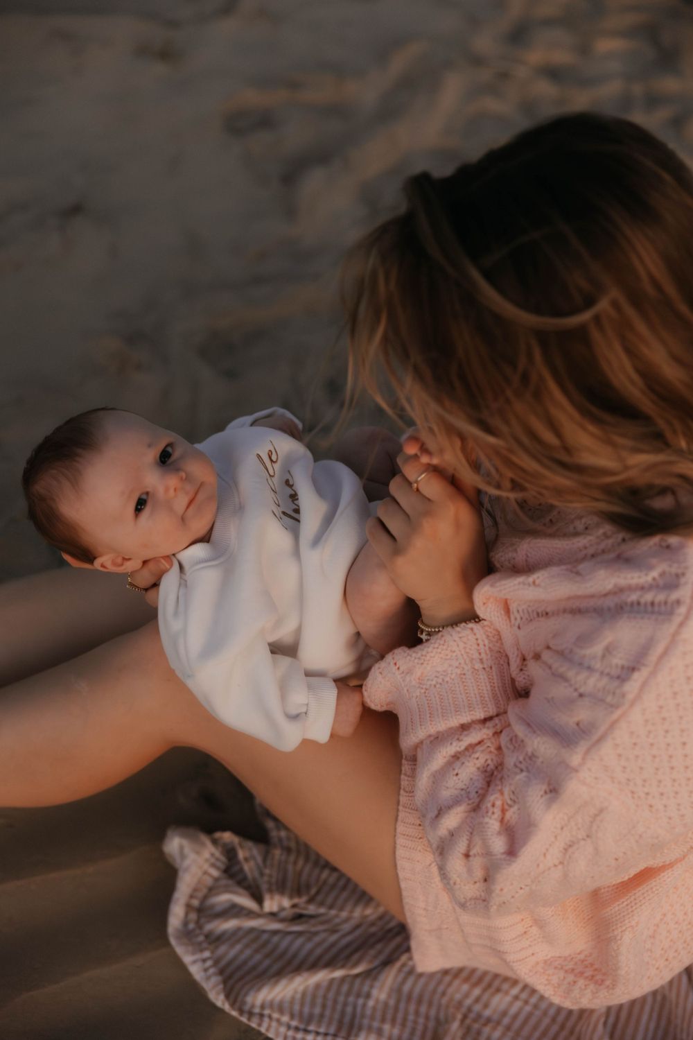 Mother in pink sweater gazing lovingly at newborn baby in white outfit on beach.