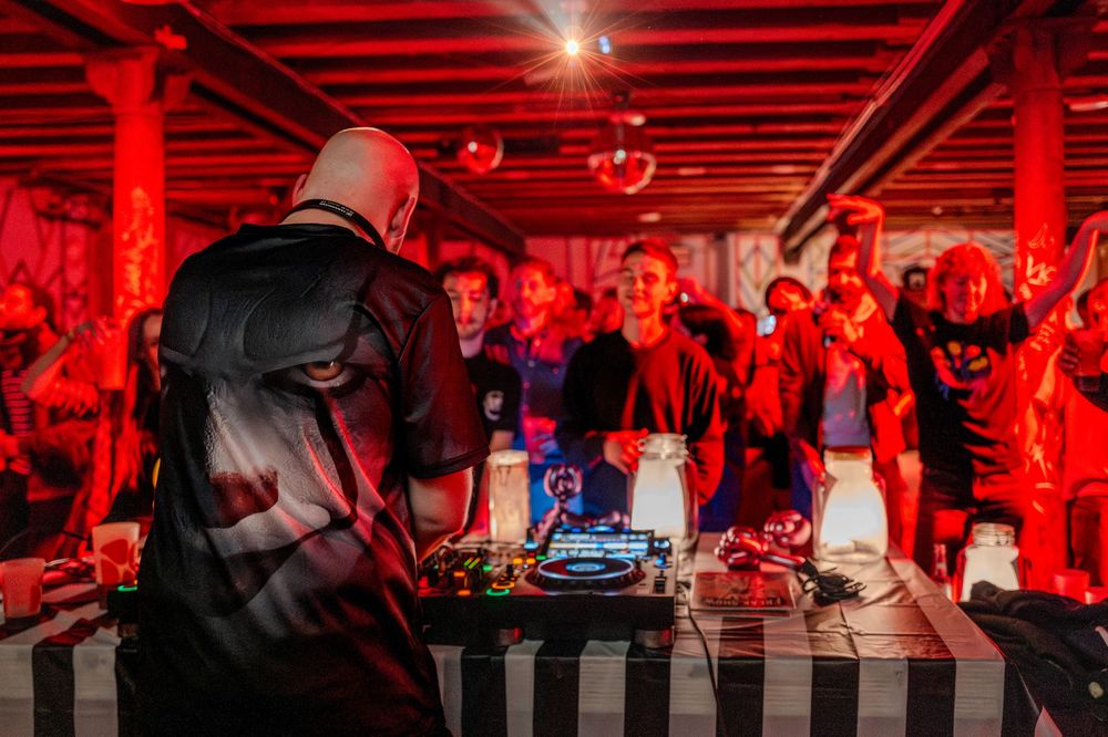 DJ performs at a nightclub bathed in dramatic red lighting while crowd dances in the background.