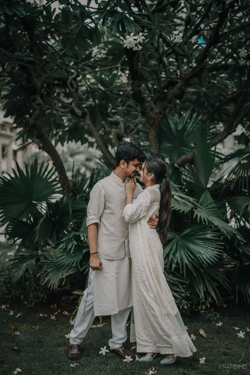Romantic couple in matching white outfits poses amid tropical foliage.