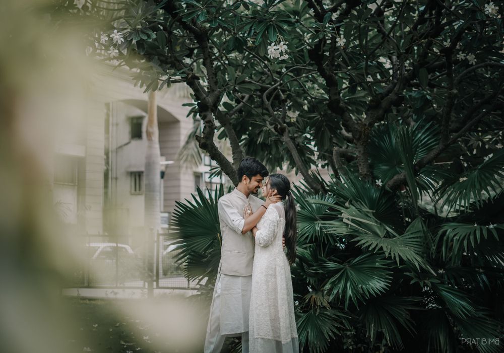 Couple in traditional white attire embraces among palm trees and garden plants.