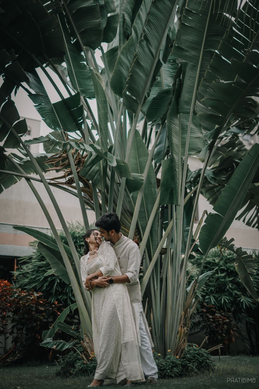 Couple poses among tall banana plants in traditional white outfits.