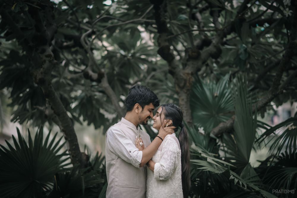 A couple in light-colored outfits embracing against a backdrop of tropical foliage.