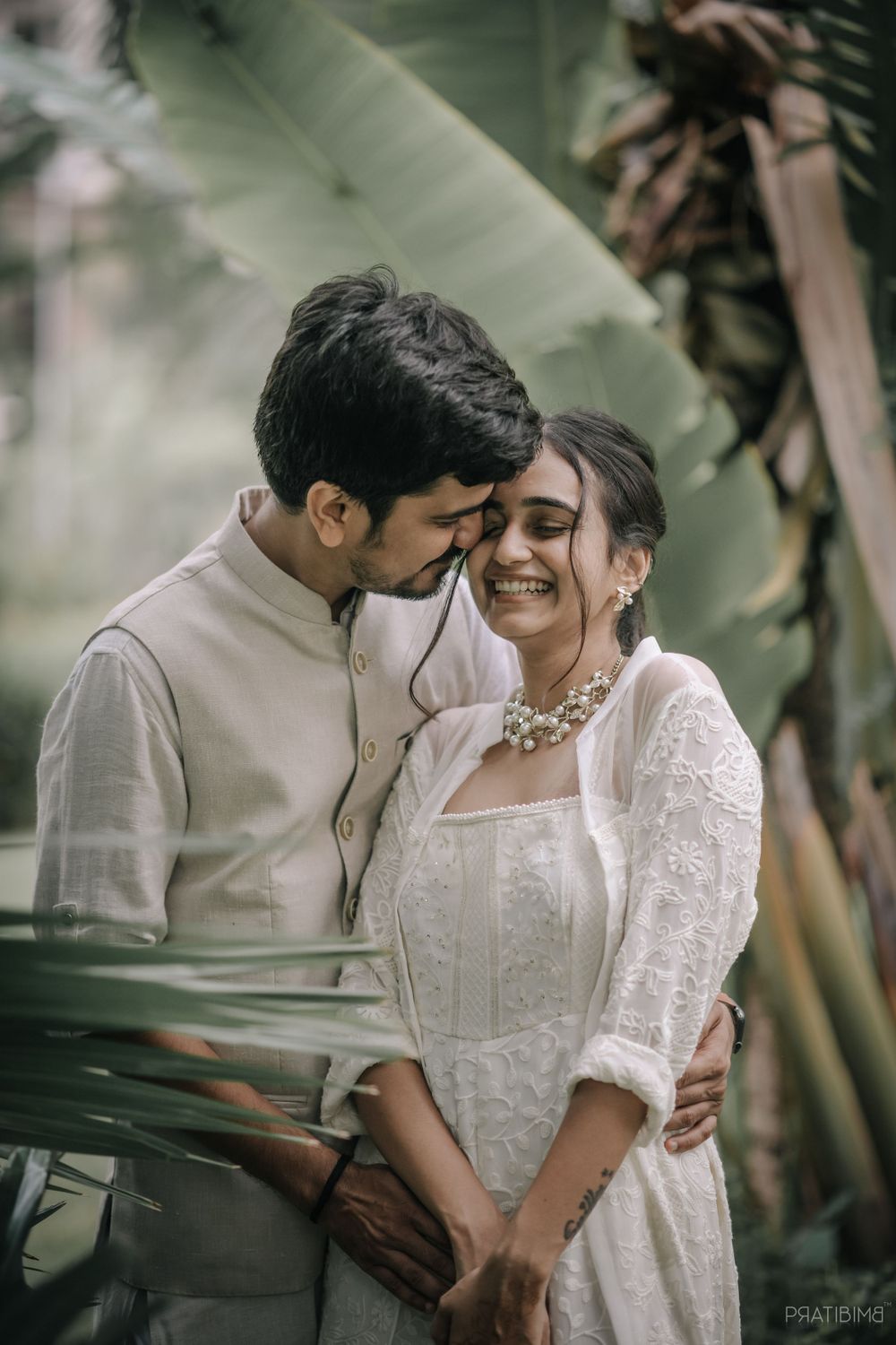 A couple sharing a joyful moment in white attire against tropical foliage.