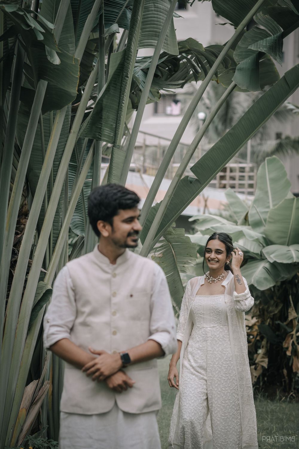 Couple in white traditional attire poses beneath banana leaves in a tropical garden setting.