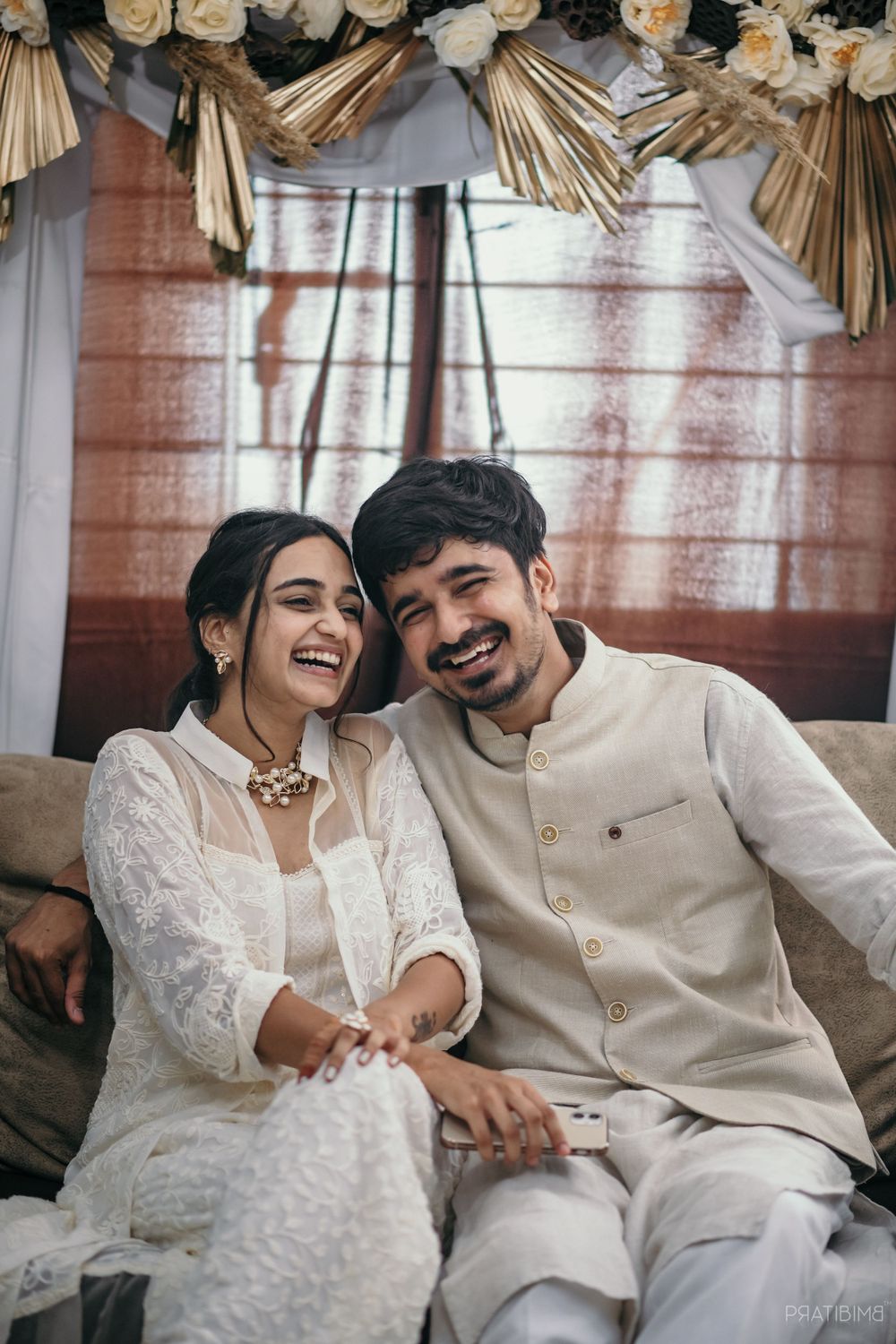 Couple in white outfits shares intimate moments on a couch with floral decorations above.