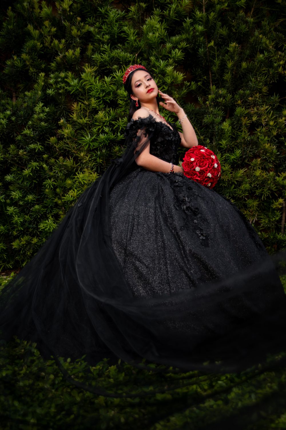 Dramatic portrait of bride in black ballgown with red bouquet against green foliage.