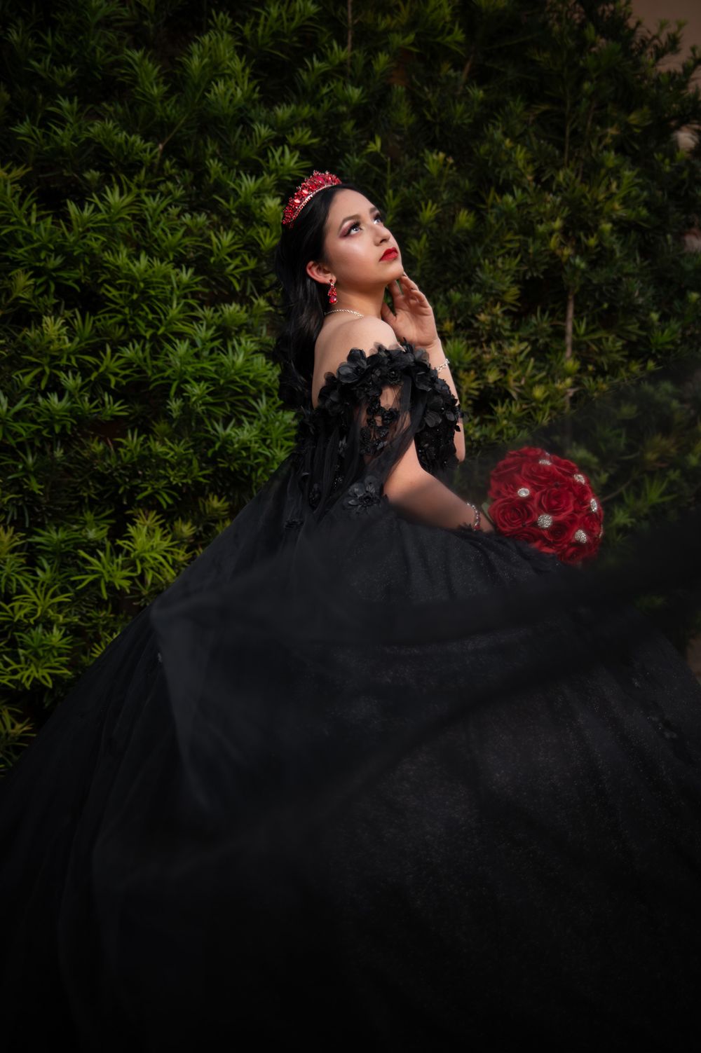 Artistic bridal portrait featuring black lace gown and red bouquet in garden setting.