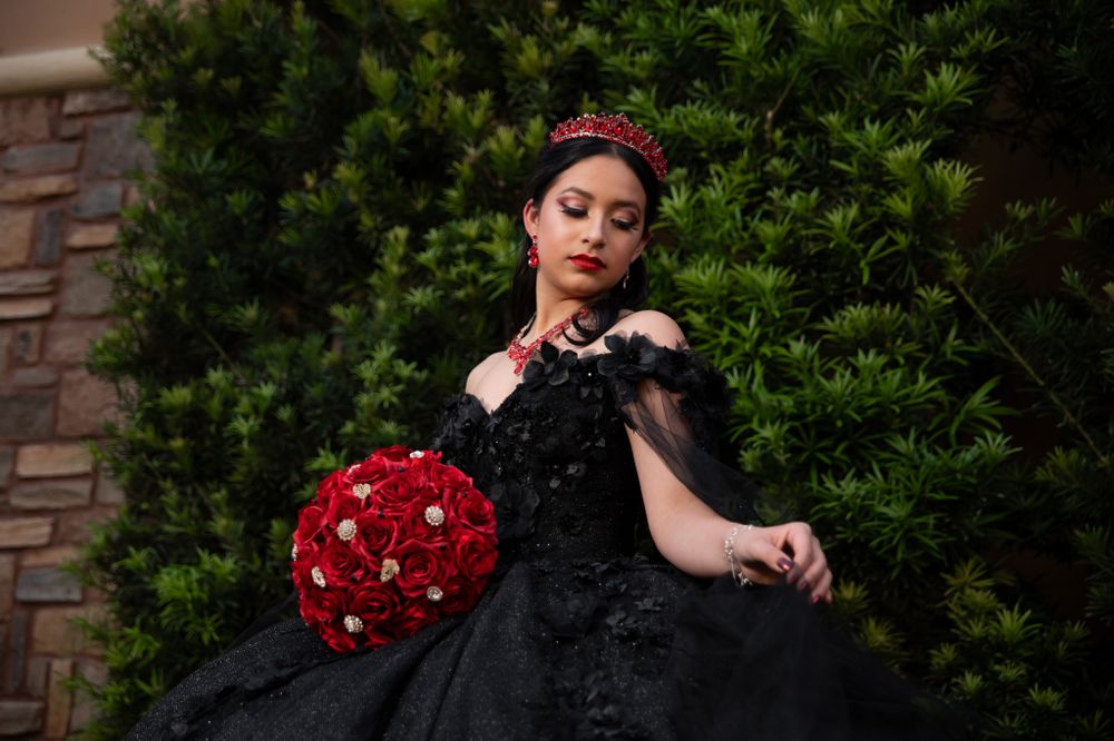 Bride poses in gothic black wedding dress with red floral bouquet against stone wall.