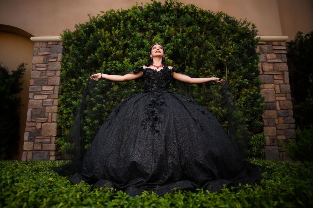 A dramatic black ball gown dress photographed against a stone archway and greenery backdrop.