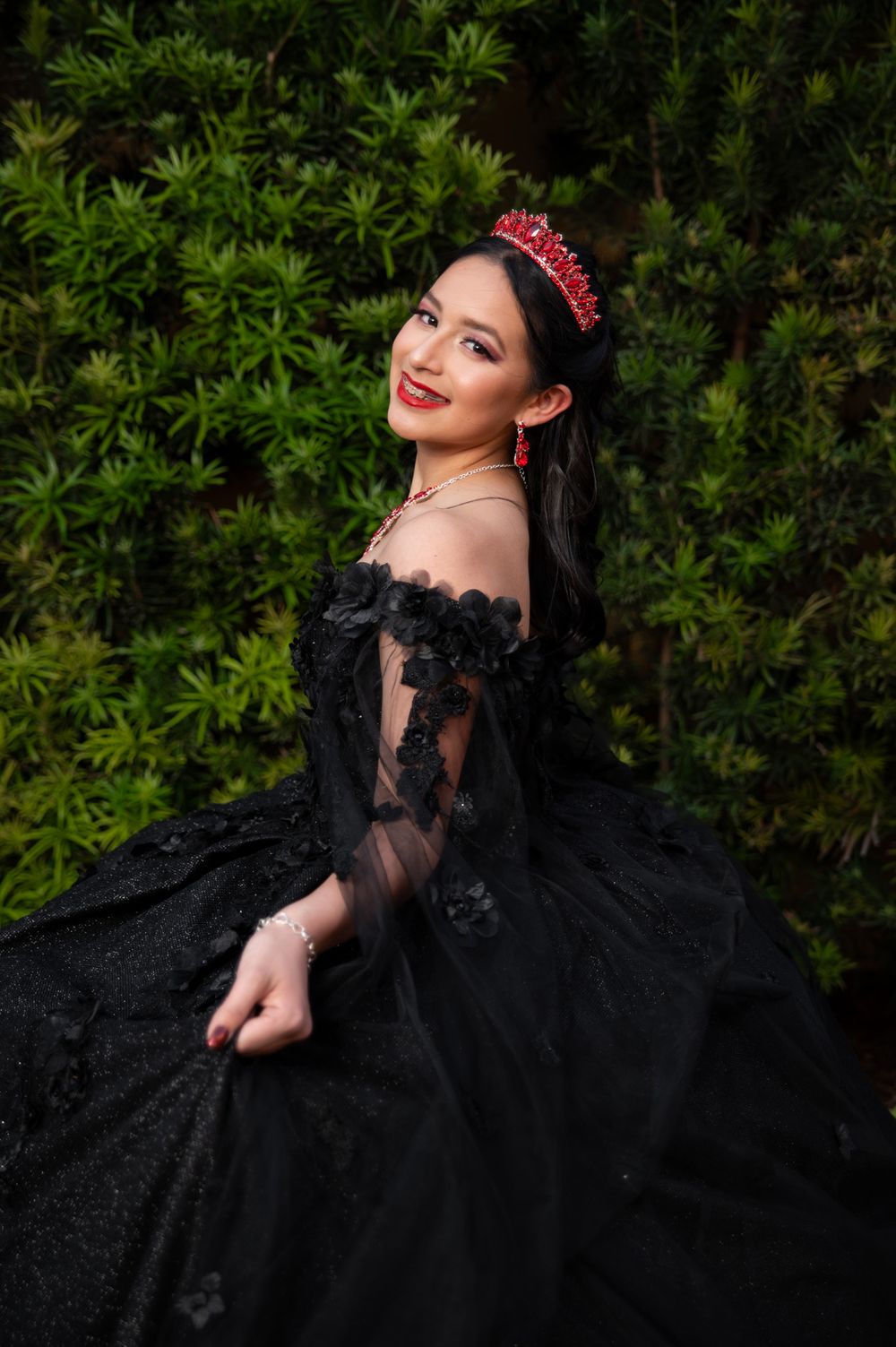 Glamorous black lace gown with red accessories against a green hedge background.