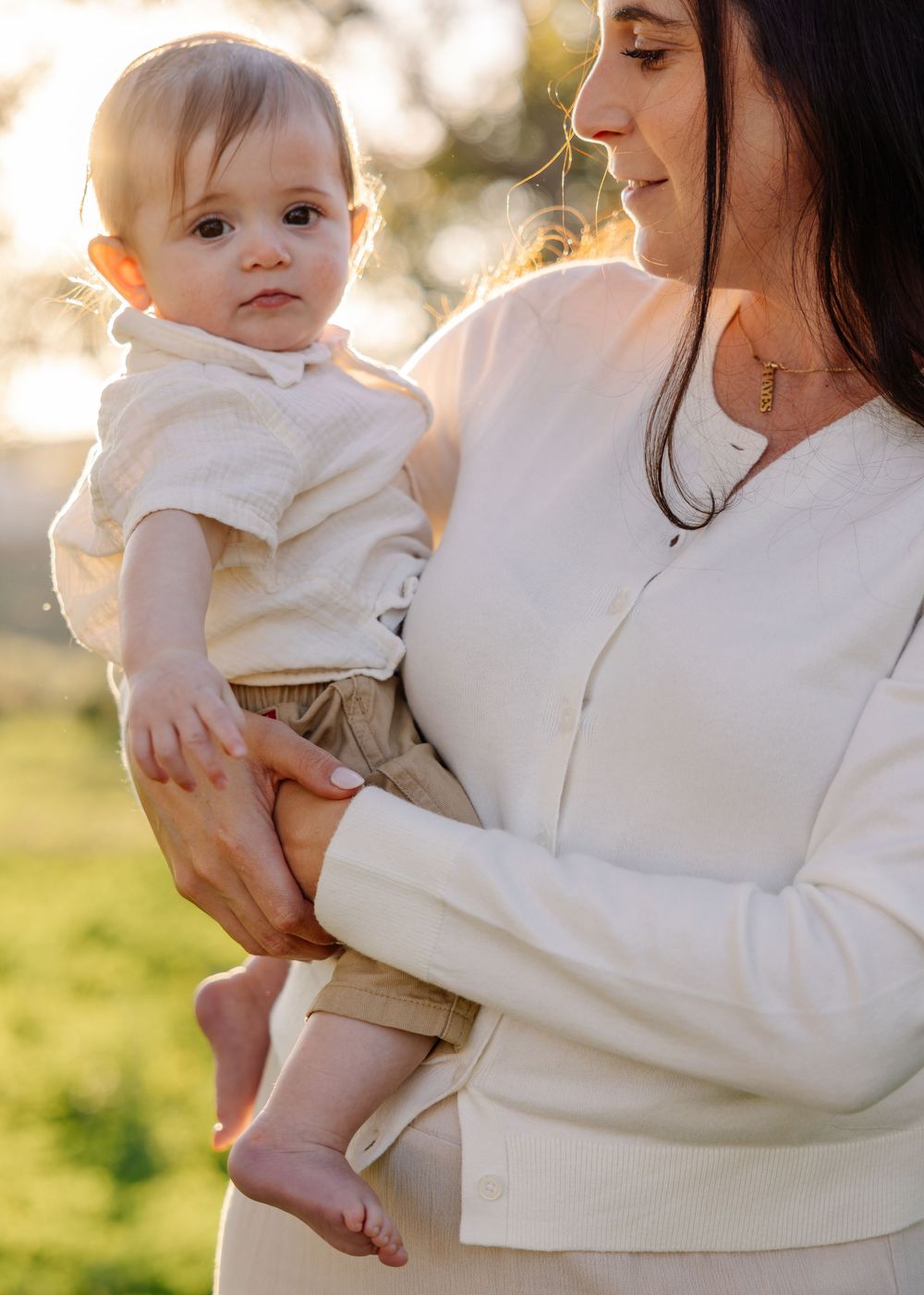 A tender moment captured in soft lighting shows a parent in white holding a baby against a sunlit outdoor background.