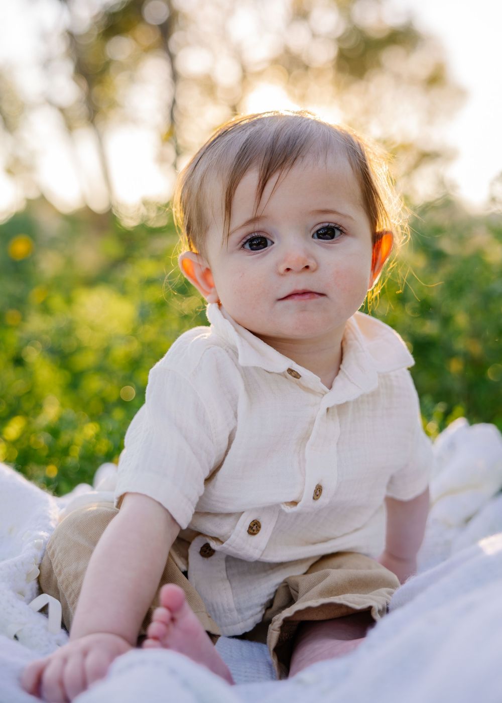 A baby in a cream-colored outfit sits on a white blanket outdoors during golden hour with greenery in the background.