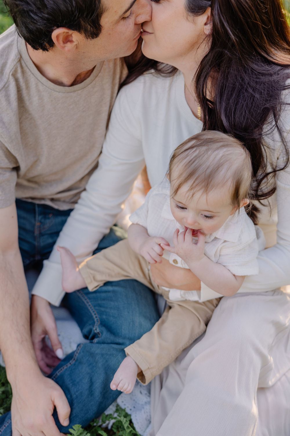 A tender family moment captured outdoors with parents and baby in coordinating neutral outfits.