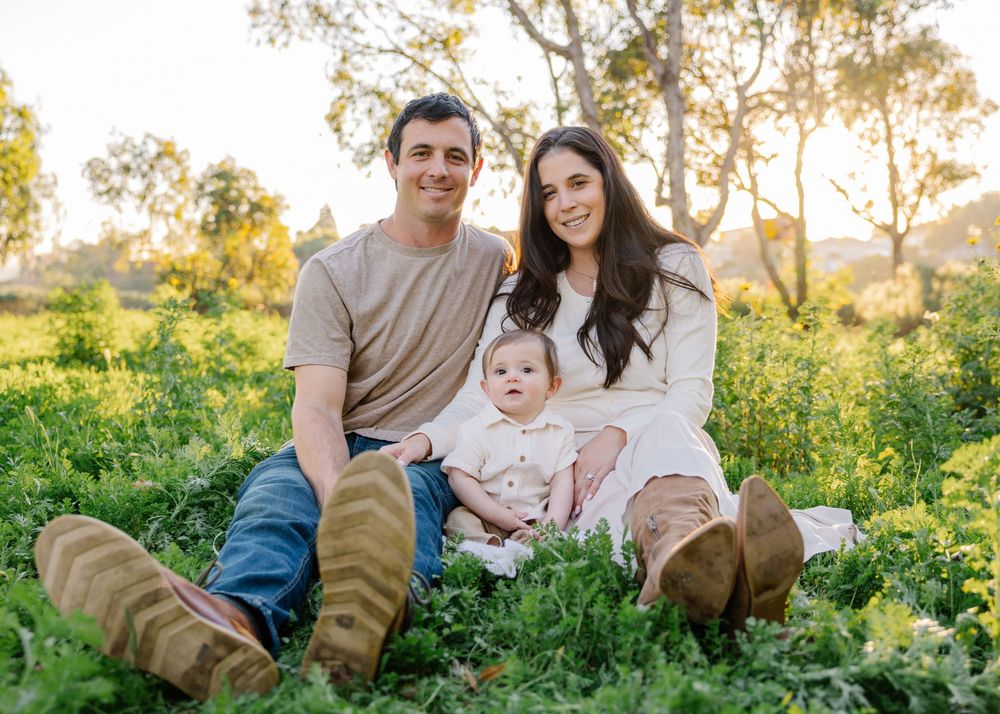 A family sits together in a sunny meadow with wildflowers and trees during golden hour.