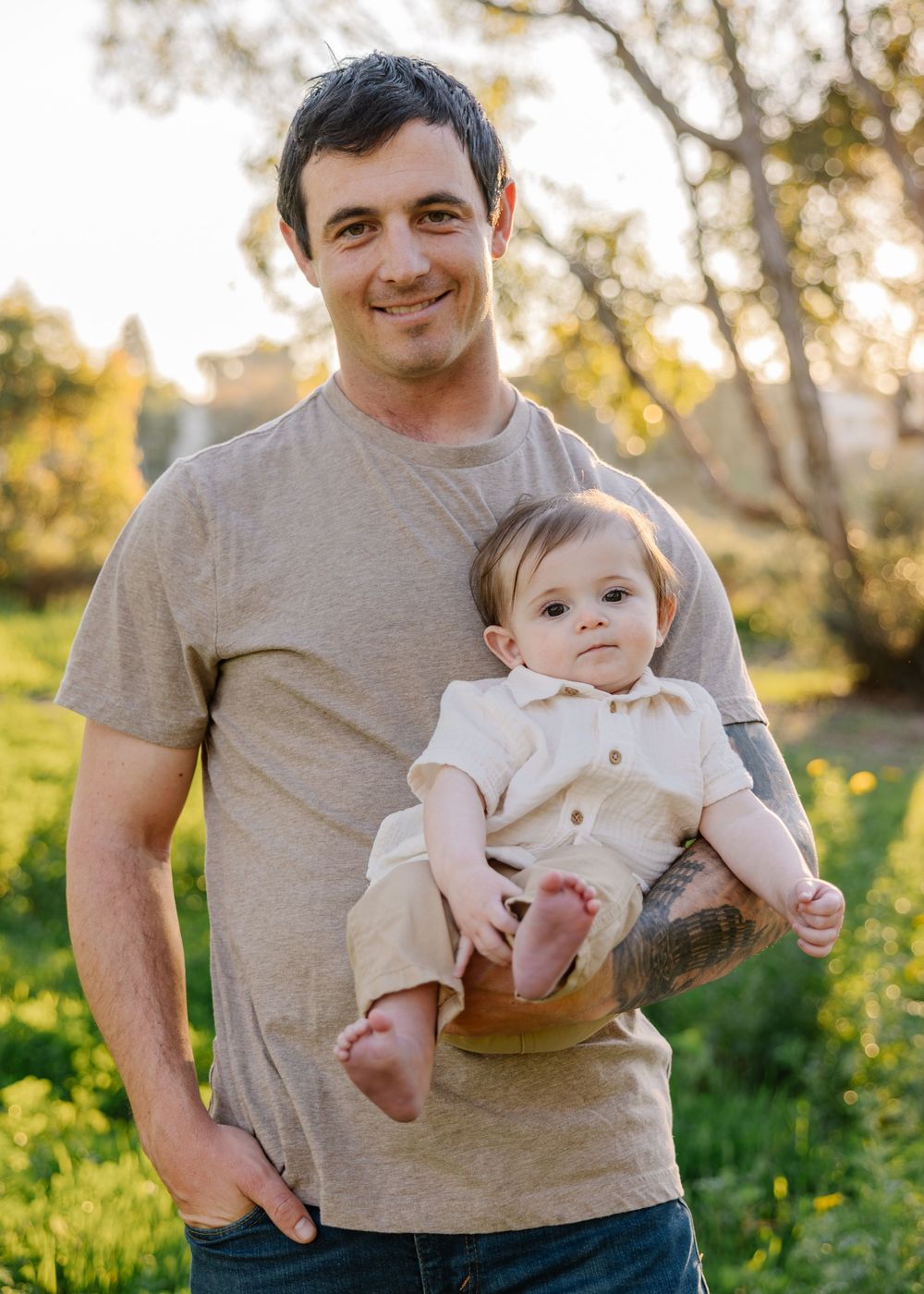 Parent in grey t-shirt holds smiling baby in light-colored outfit against natural outdoor backdrop.
