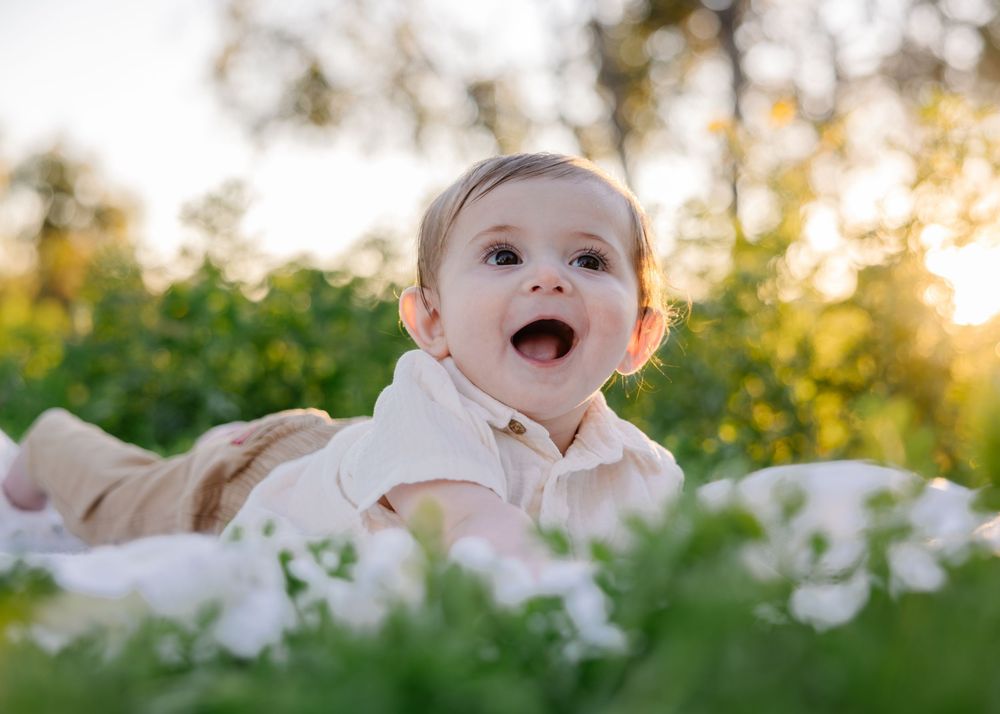 Baby lying in soft white blankets outdoors surrounded by wildflowers at sunset.