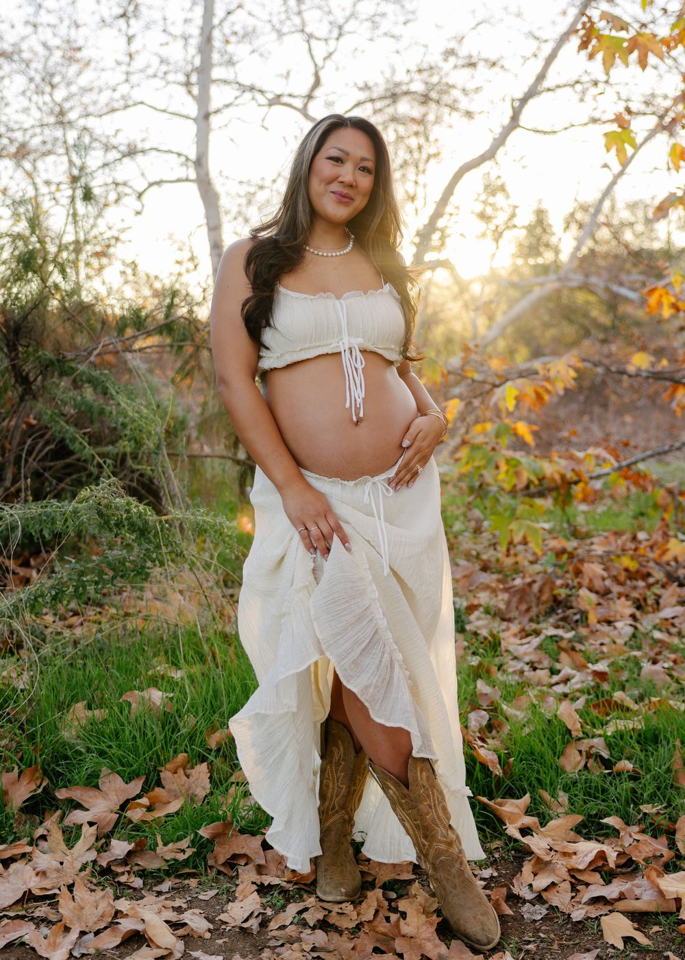 A maternity photoshoot in a natural fall setting with autumn leaves and golden sunlight filtering through trees.