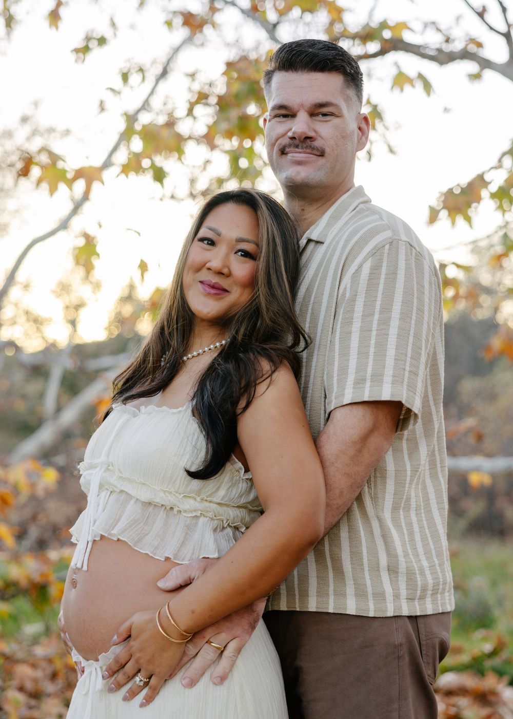 A couple poses for maternity photos in a scenic outdoor setting with autumn leaves in the background.