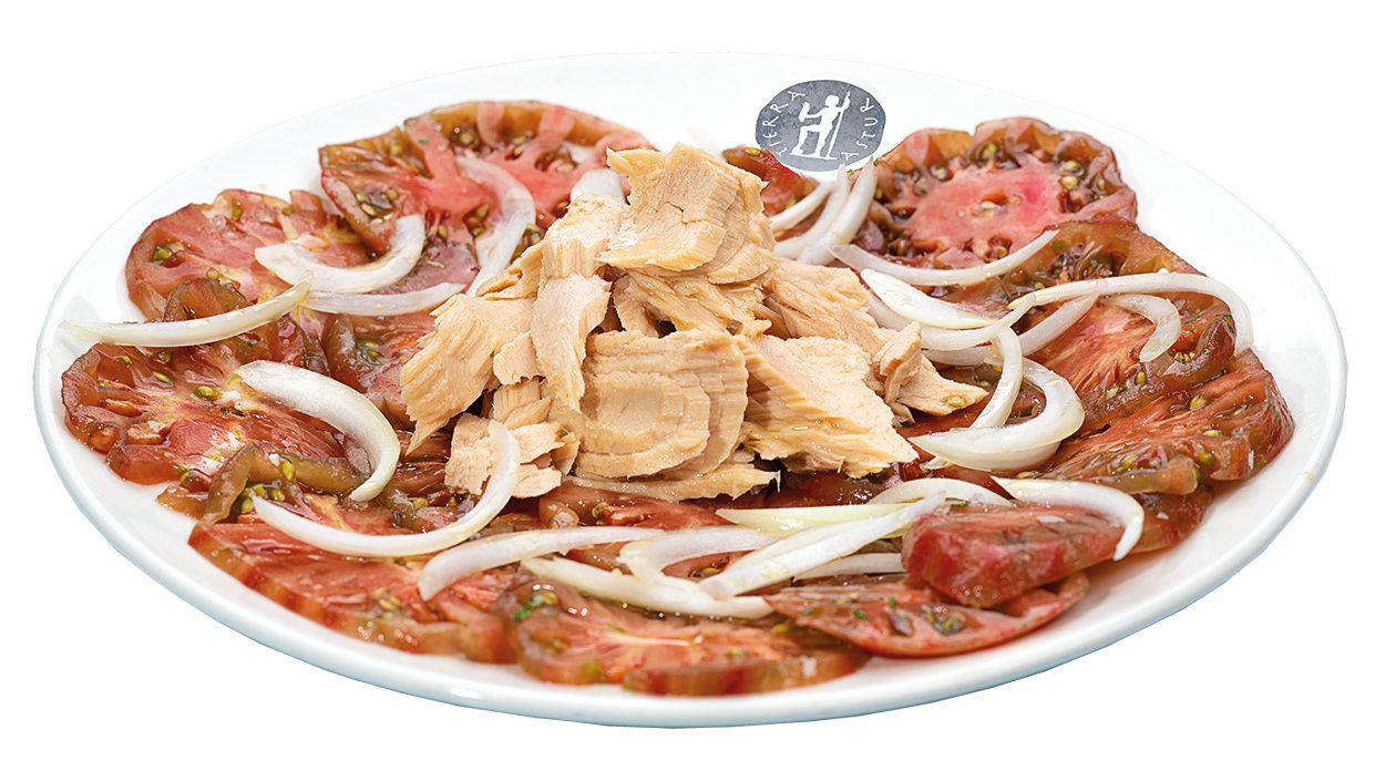 Un plato redondo de tomate en rodajas finas adornado con aros de cebolla y bonito. Tierra Astur