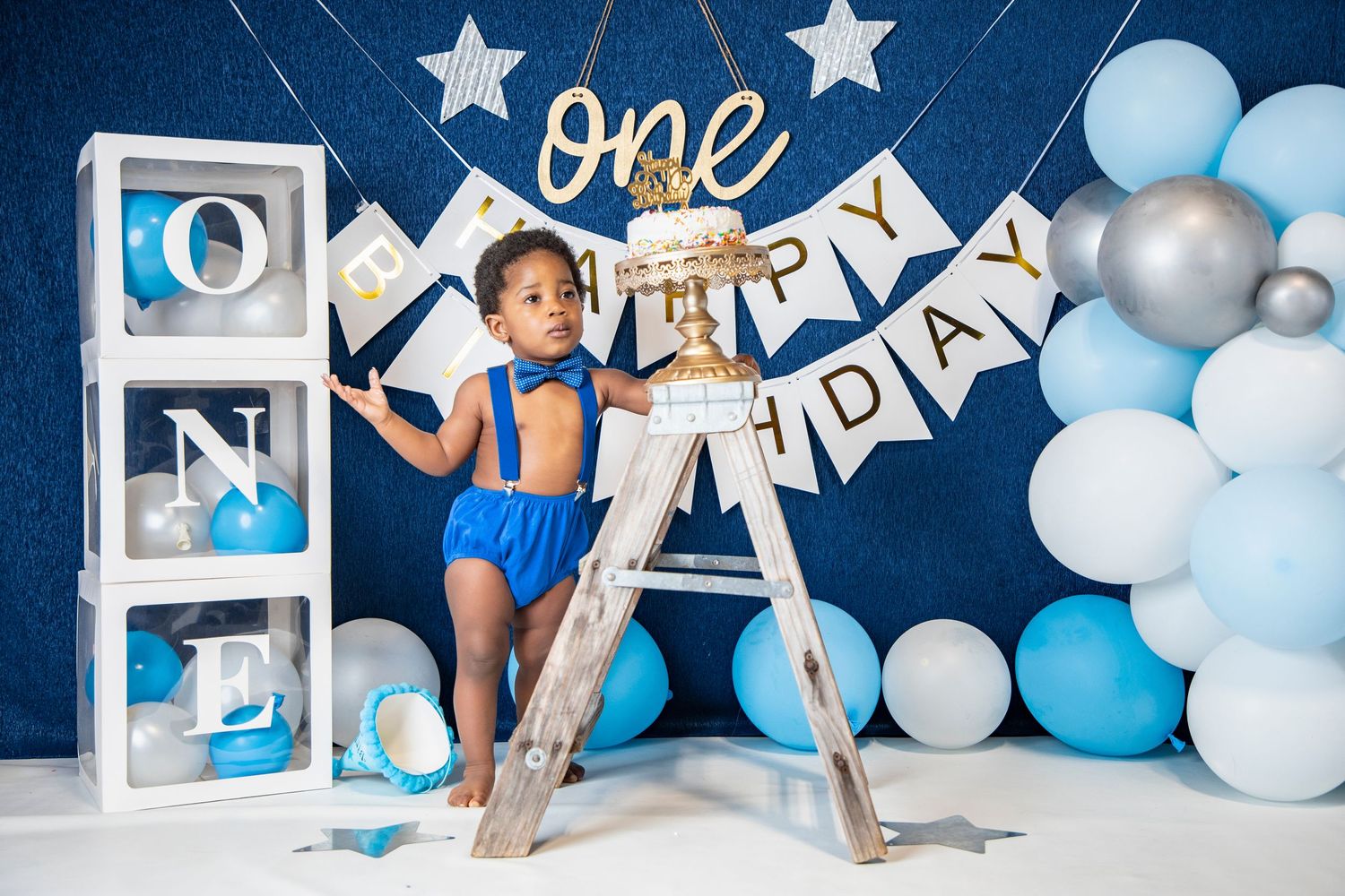 First birthday party scene with blue and white balloons, stars, and decorative frames.