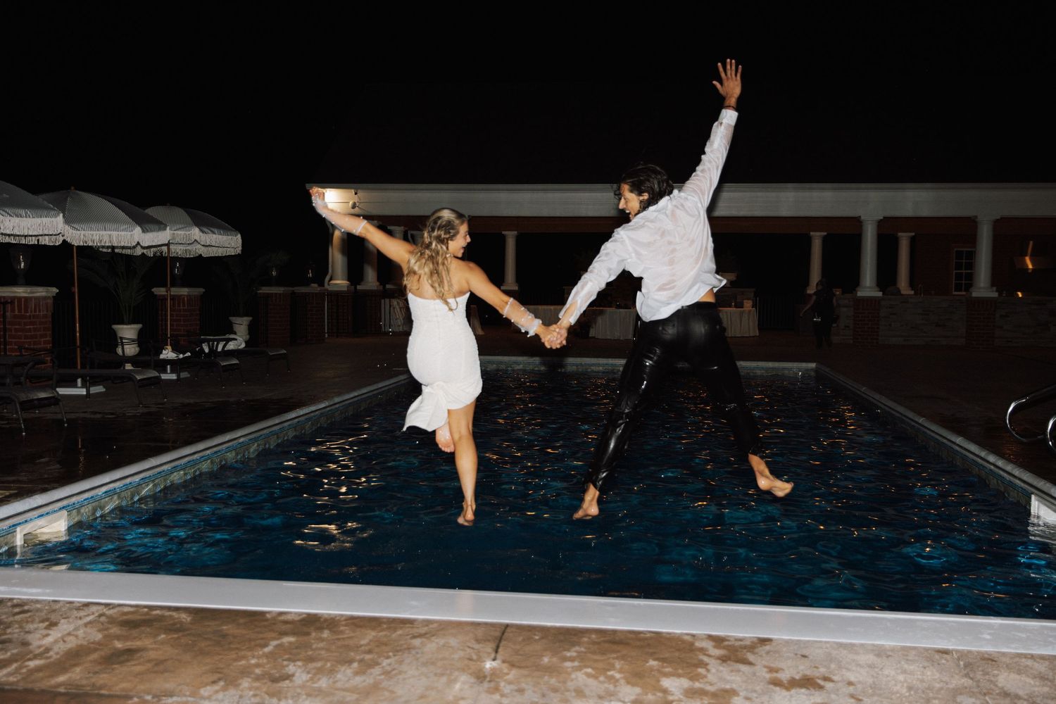 A celebratory couple in formal white attire jumps into a swimming pool at night during what appears to be a special event.