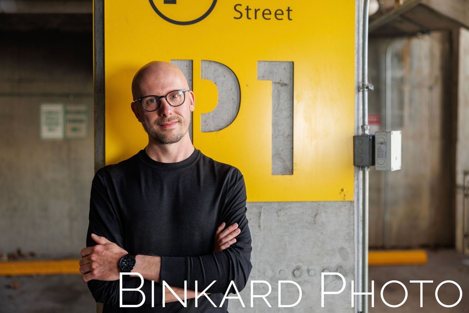 Professional in black sweater standing with crossed arms against industrial yellow and gray wall with numbers.