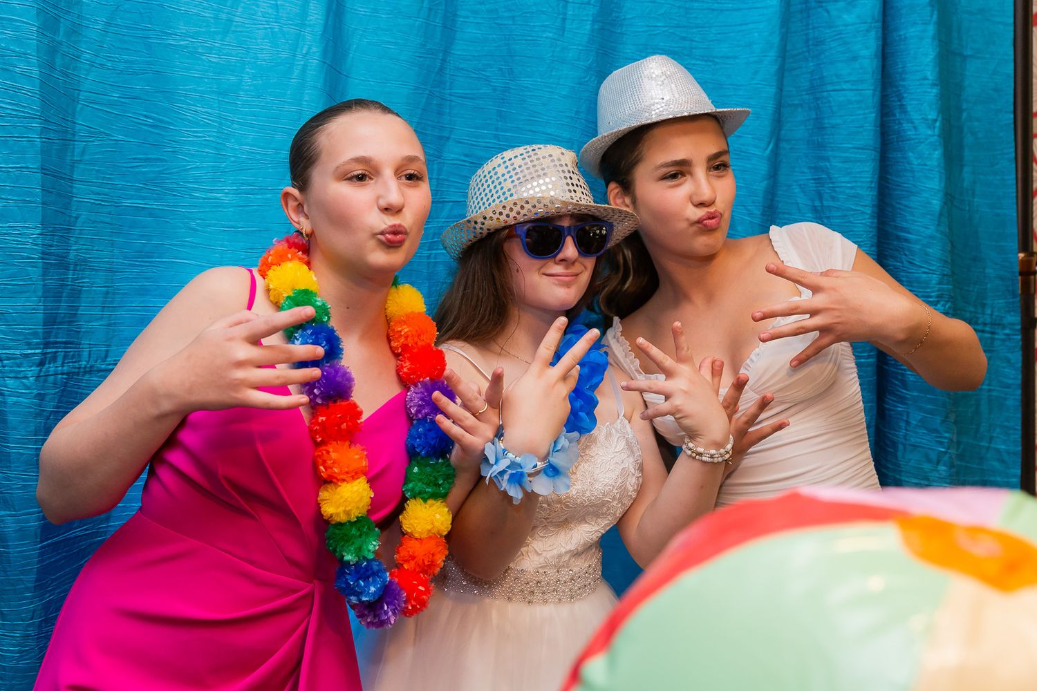 Three friends pose with colorful party props and leis against a turquoise backdrop at a photo booth.