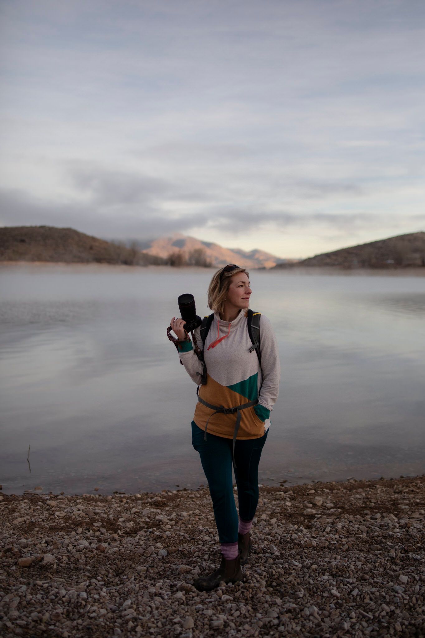 Ali of Lumenali Photography holds her camera while standing on a shoreline with foggy mountains in the background. Ontario, Canada.