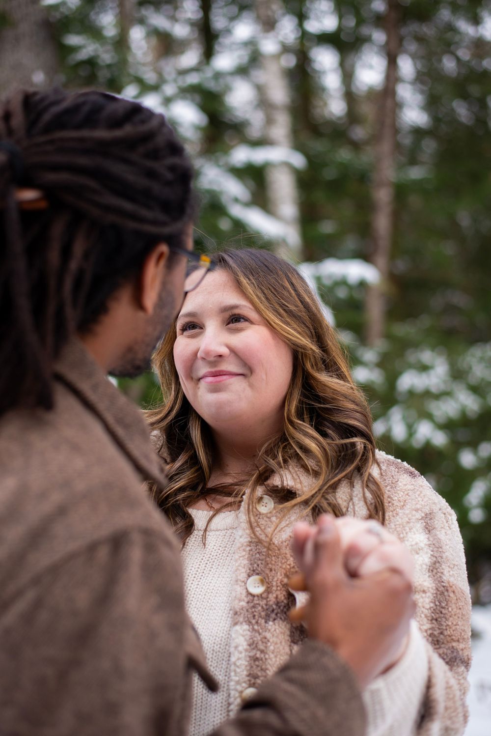 A couple shares an intimate moment together in the snowy woods wearing cozy winter sweaters.