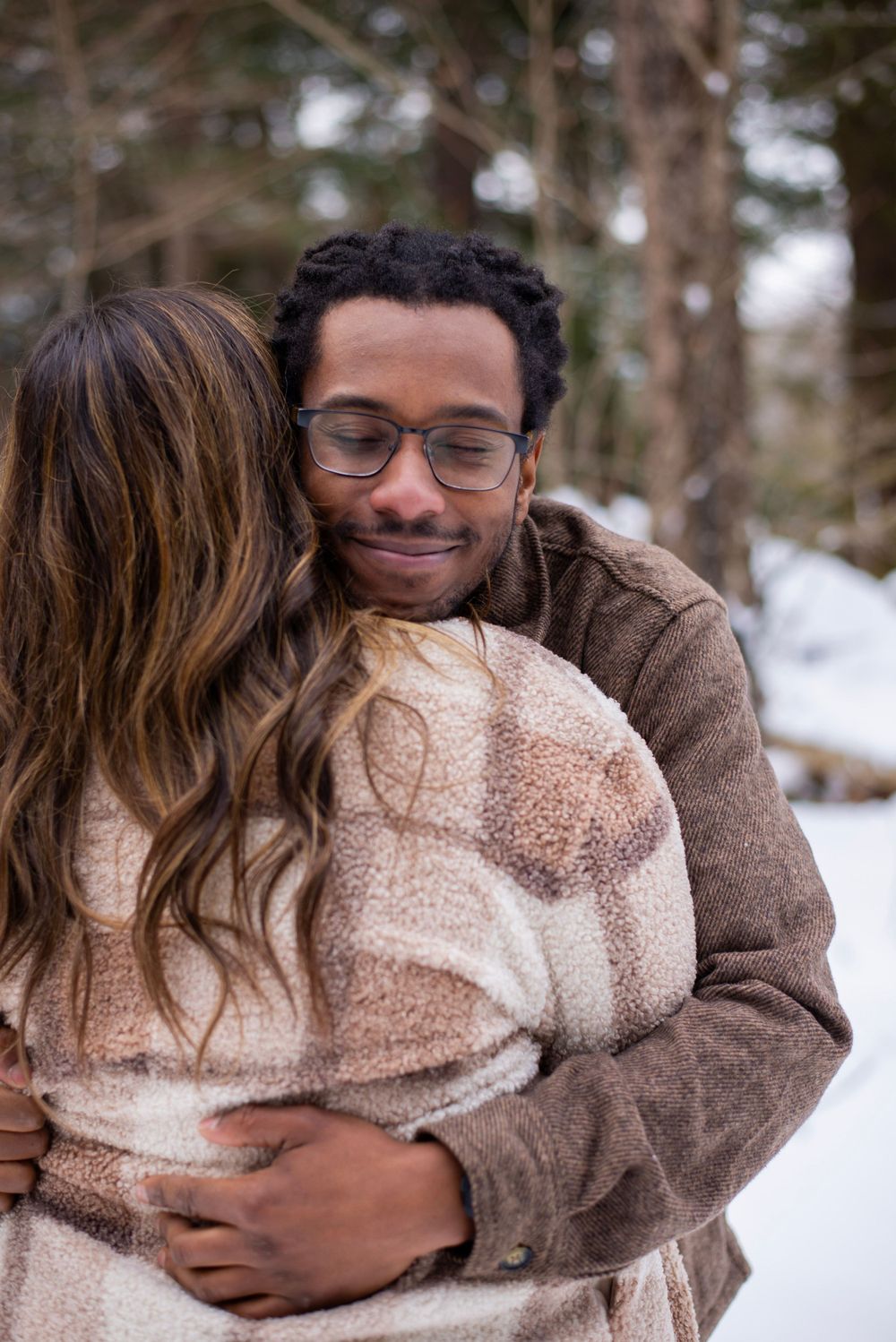 A couple shares a tender embrace while wearing cozy sweaters in a snowy winter forest setting.